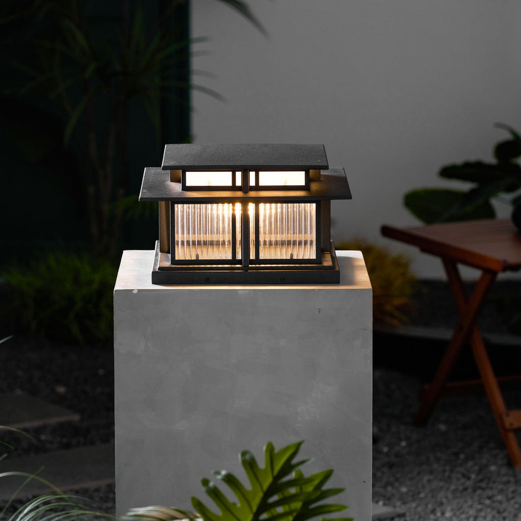 A different angle of this lit Clérachic pillar light on its concrete plinth, framed by dark garden greenery.