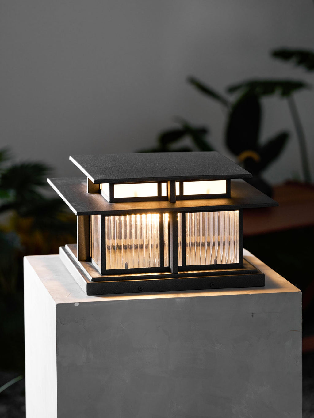 Indoors, this lit Clérachic pillar light sits on a pale pedestal, its frosted glass panels glowing against a dark backdrop.