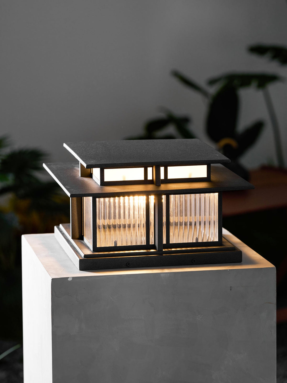 Indoors, this lit Clérachic pillar light sits on a pale pedestal, its frosted glass panels glowing against a dark backdrop.