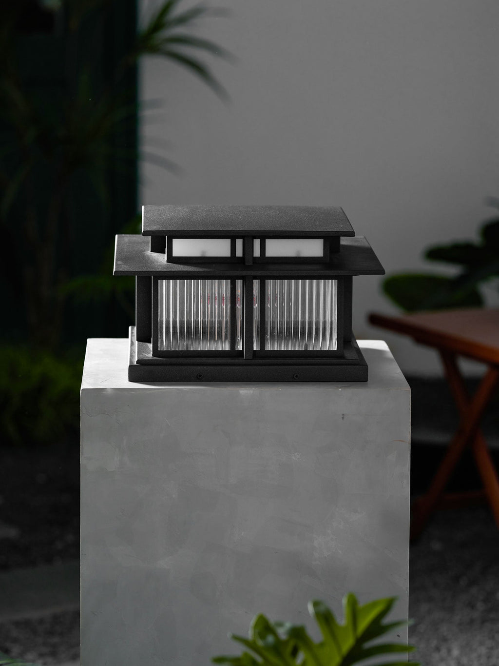 From a different angle, this lit Clérachic pillar light glows atop a stone plinth beside potted greenery at night.