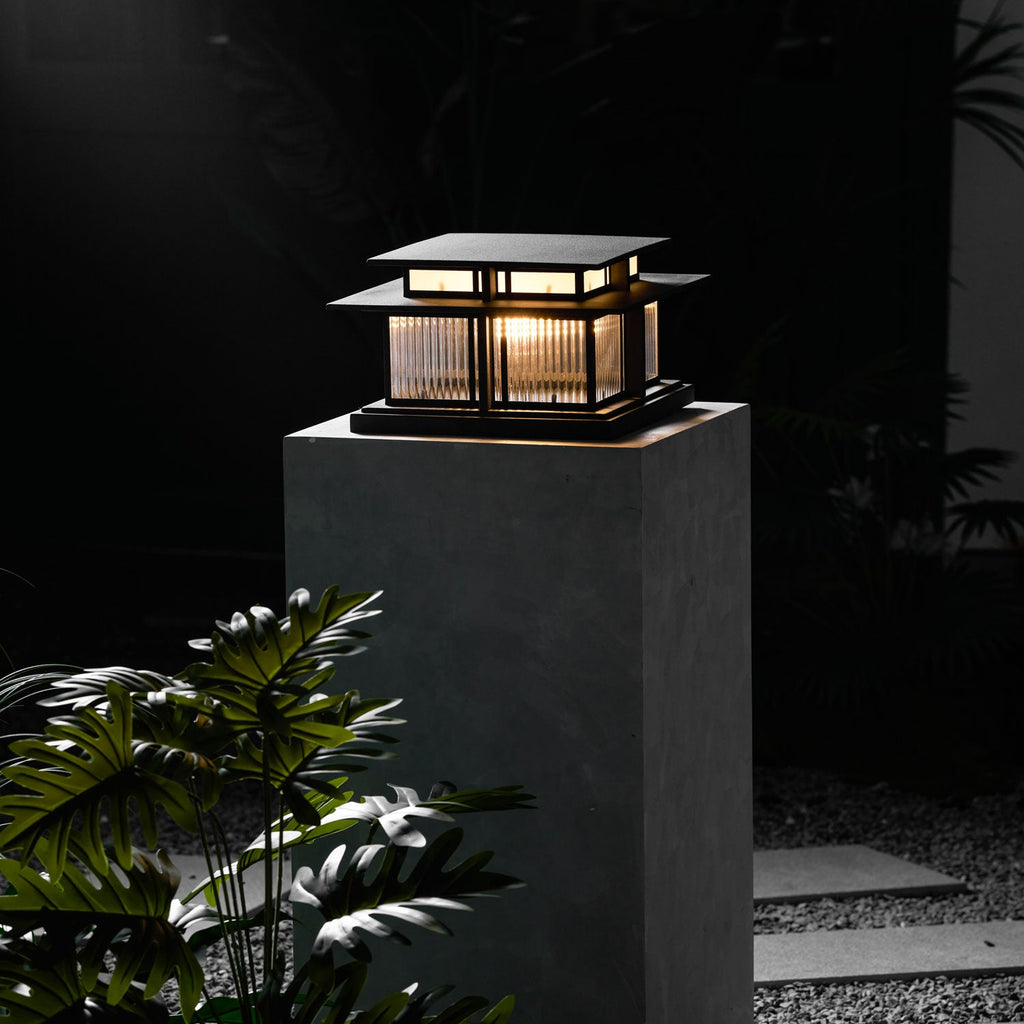 A taller framing shows this lit Clérachic pillar light glowing atop its stone plinth among dark garden foliage.
