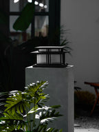 In a vertical composition, this lit Clérachic pillar light glows atop a stone plinth beside a window and greenery.