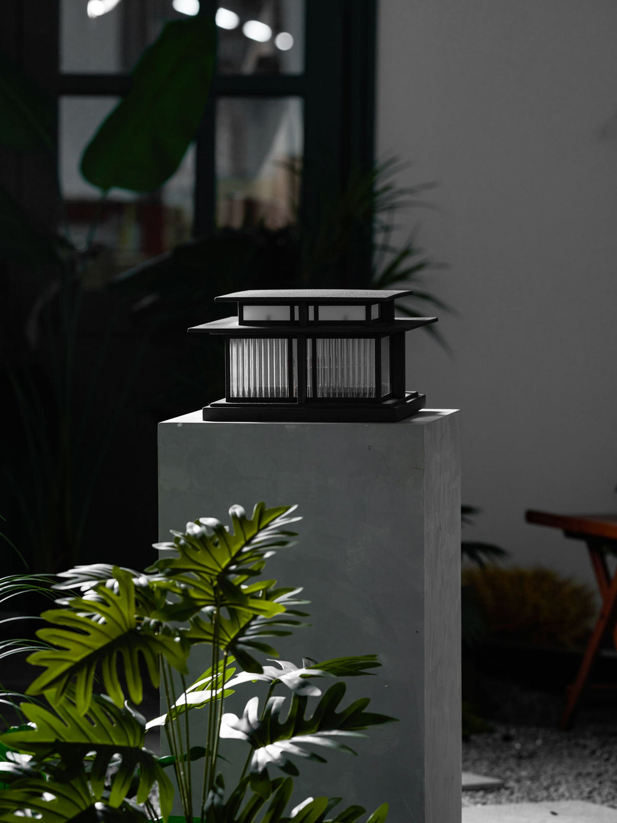 In a vertical composition, this lit Clérachic pillar light glows atop a stone plinth beside a window and greenery.