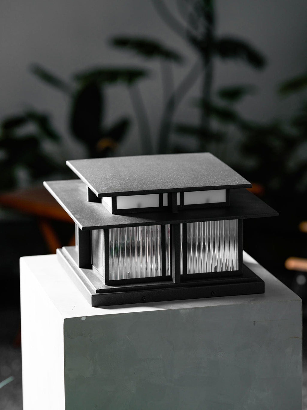 A close angled view of this lit Clérachic pillar light on its stone plinth, dark foliage surrounding it.