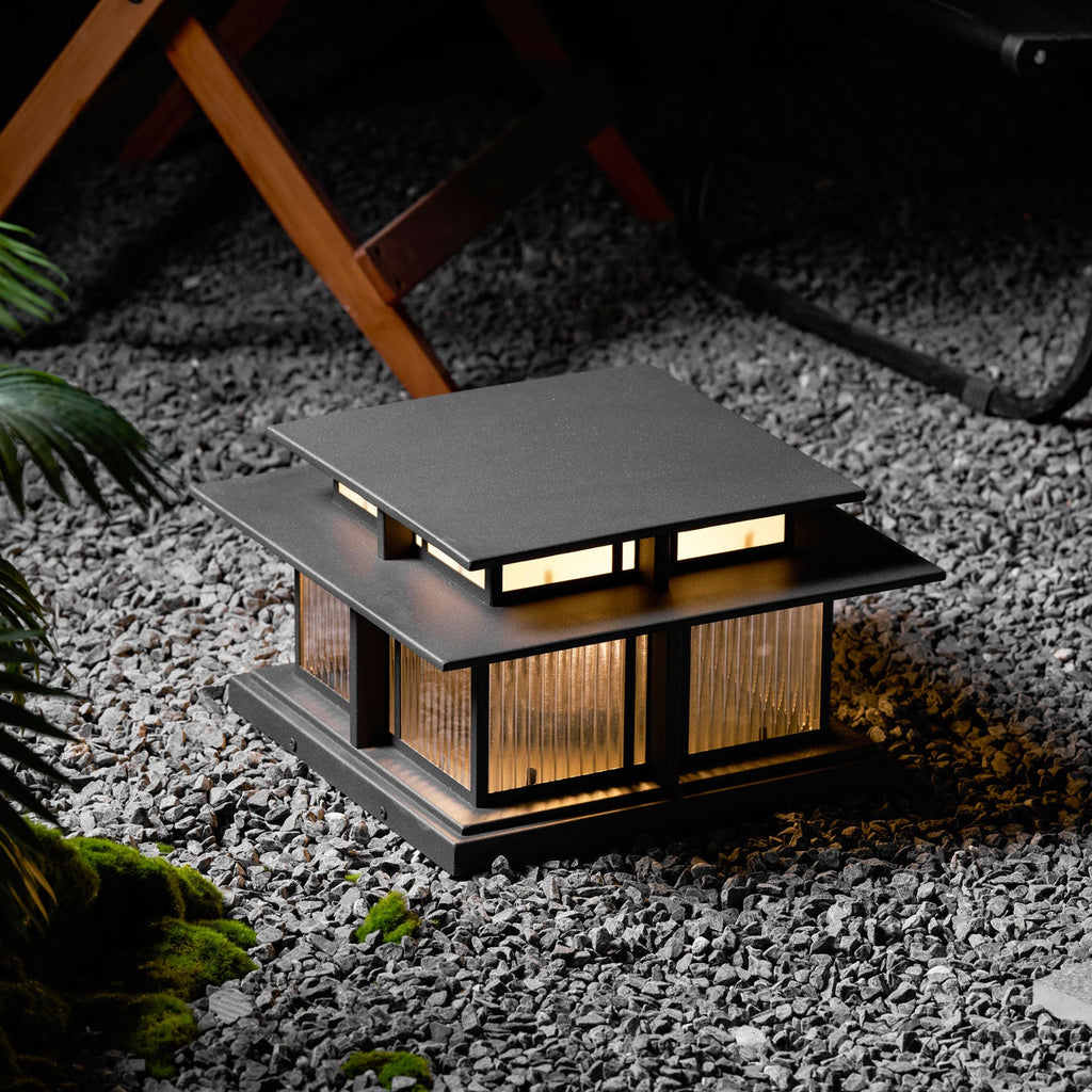 Set on pale gravel, this lit Clérachic pillar light glows beside foliage, a wooden chair blurred in the background.