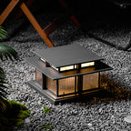 Set on pale gravel, this lit Clérachic pillar light glows beside foliage, a wooden chair blurred in the background.