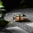 This lit Clérachic pillar light stands on gravel beside a stepping-stone path, plants softly lit around its base.