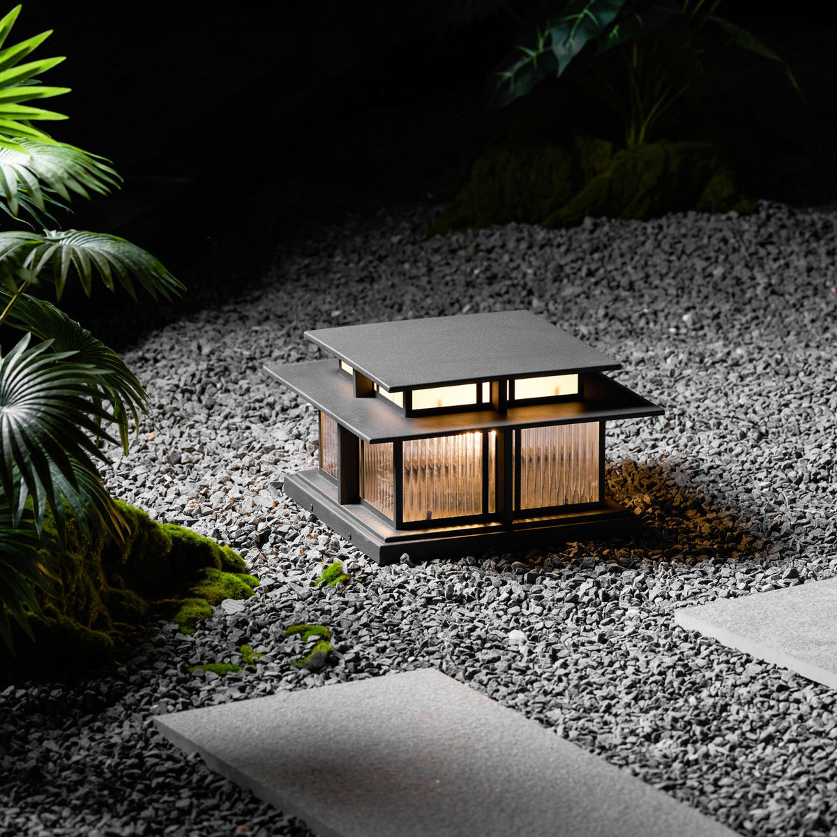 This lit Clérachic pillar light stands on gravel beside a stepping-stone path, plants softly lit around its base.