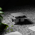 Another gravel garden view of this lit Clérachic pillar light, its warm glow set against dark, textured stone.