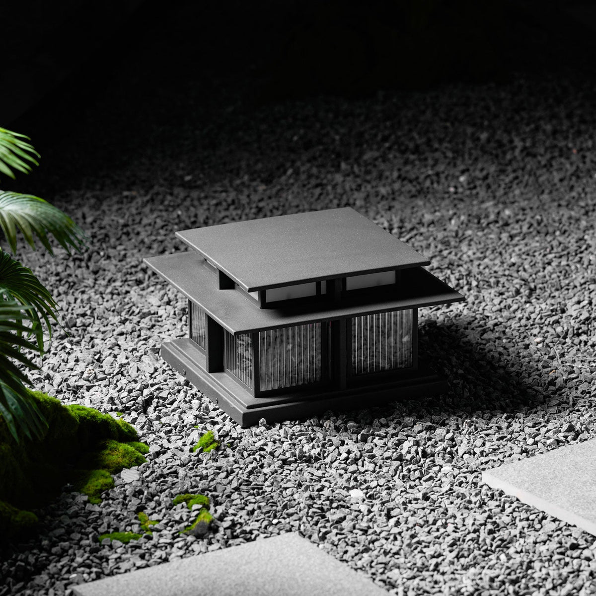 Another gravel garden view of this lit Clérachic pillar light, its warm glow set against dark, textured stone.