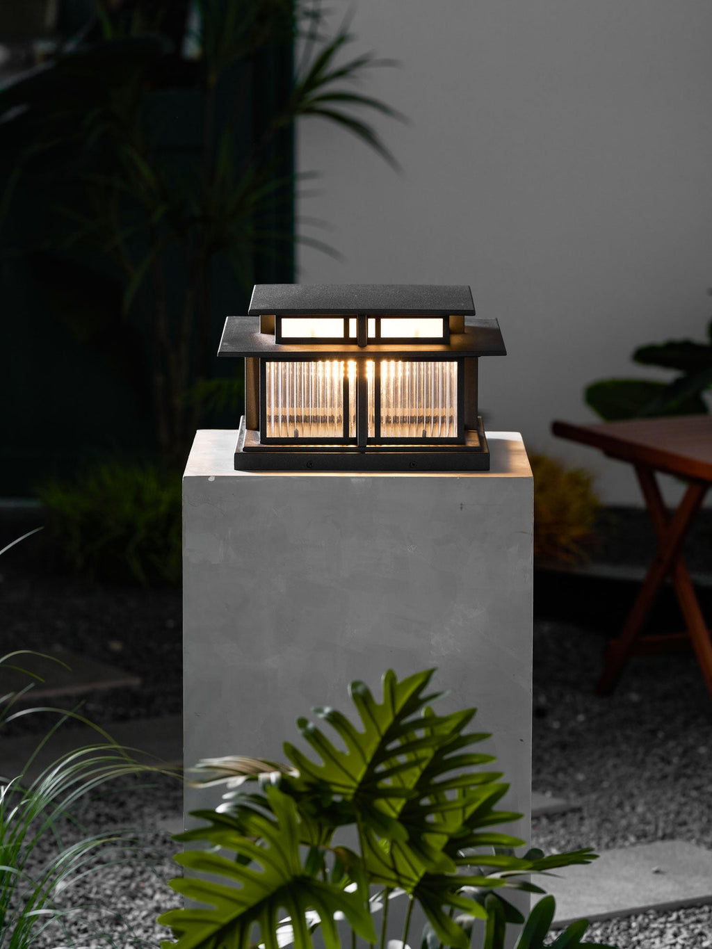 A tall concrete plinth raises this lit Clérachic pillar light in a night garden, potted plants nearby.