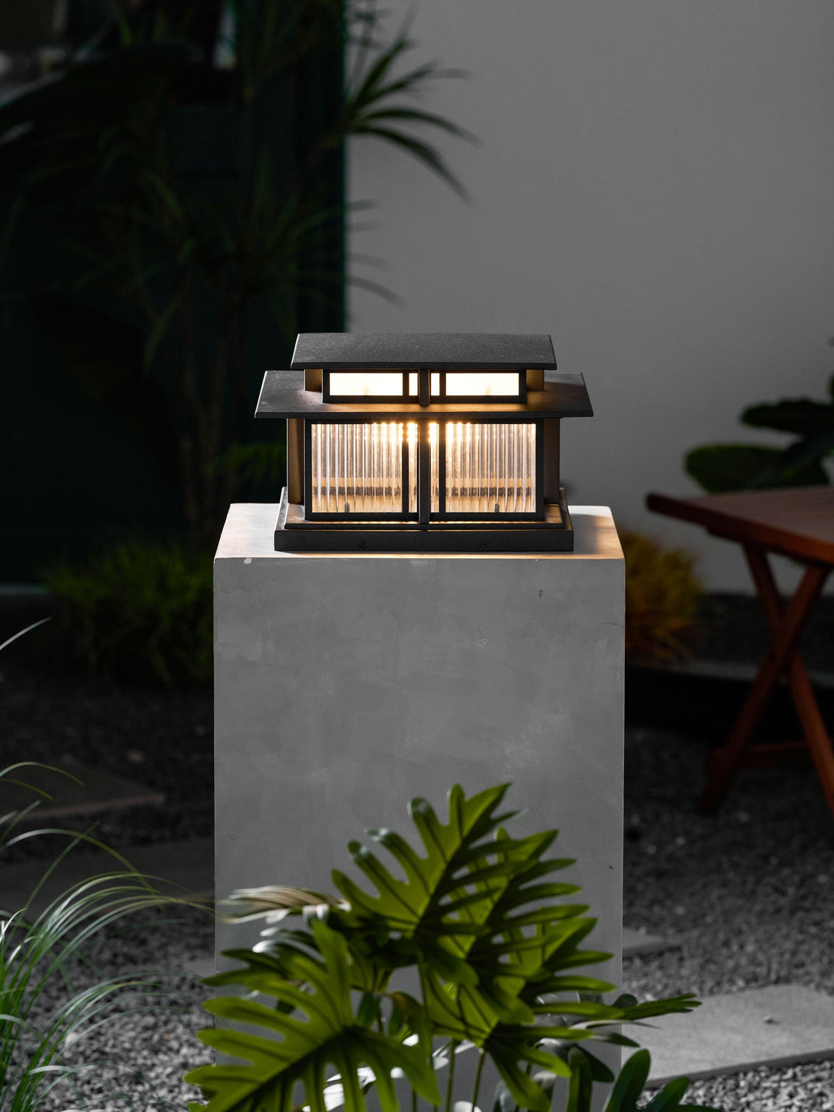 A tall concrete plinth raises this lit Clérachic pillar light in a night garden, potted plants nearby.