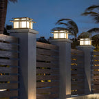 Three lit Clérachic pillar lights sit atop posts along a wooden slat fence at dusk, palm fronds overhead.