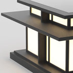 Angled view of this lit Clérachic pillar light, warm light spilling through its frosted glass panels against a textured grey backdrop.
