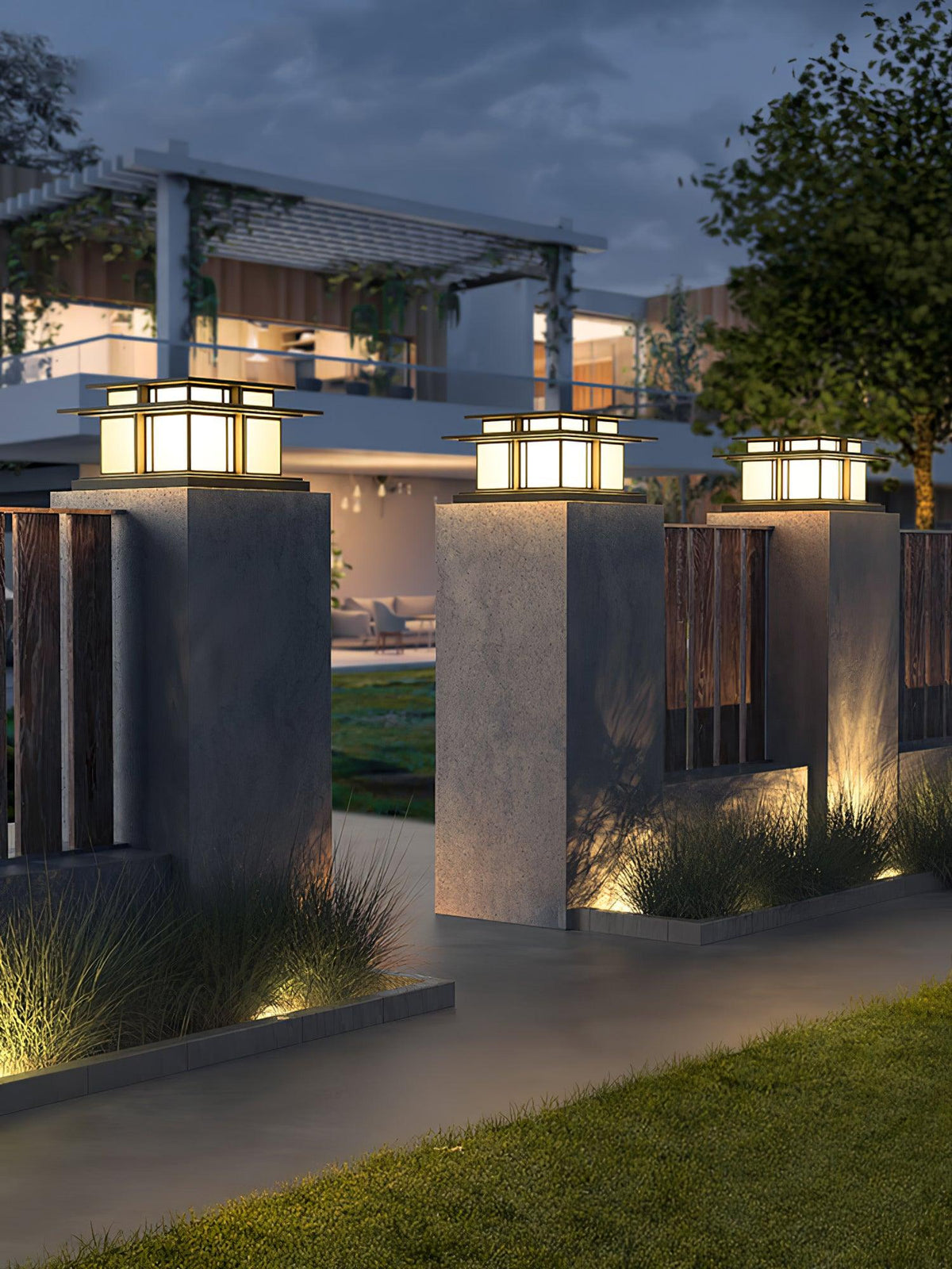 Three lit Clérachic pillar lights line a garden path beneath a pergola, grass and shrubs at their base.
