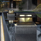 A pair of lit Clérachic pillar lights stands beside a reflecting pool, the nearer one raised on a stone plinth.