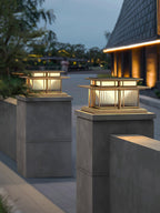 In a paved courtyard, two lit Clérachic pillar lights top concrete posts beneath a sloped roofline at dusk.