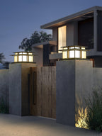 Two lit Clérachic pillar lights flank a wood-panelled gate between concrete posts, a modern house behind.