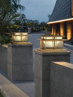 Two lit lanterns with a gold-toned frame stand atop stone posts at dusk, a different finish from this Clérachic pillar light's black frame.