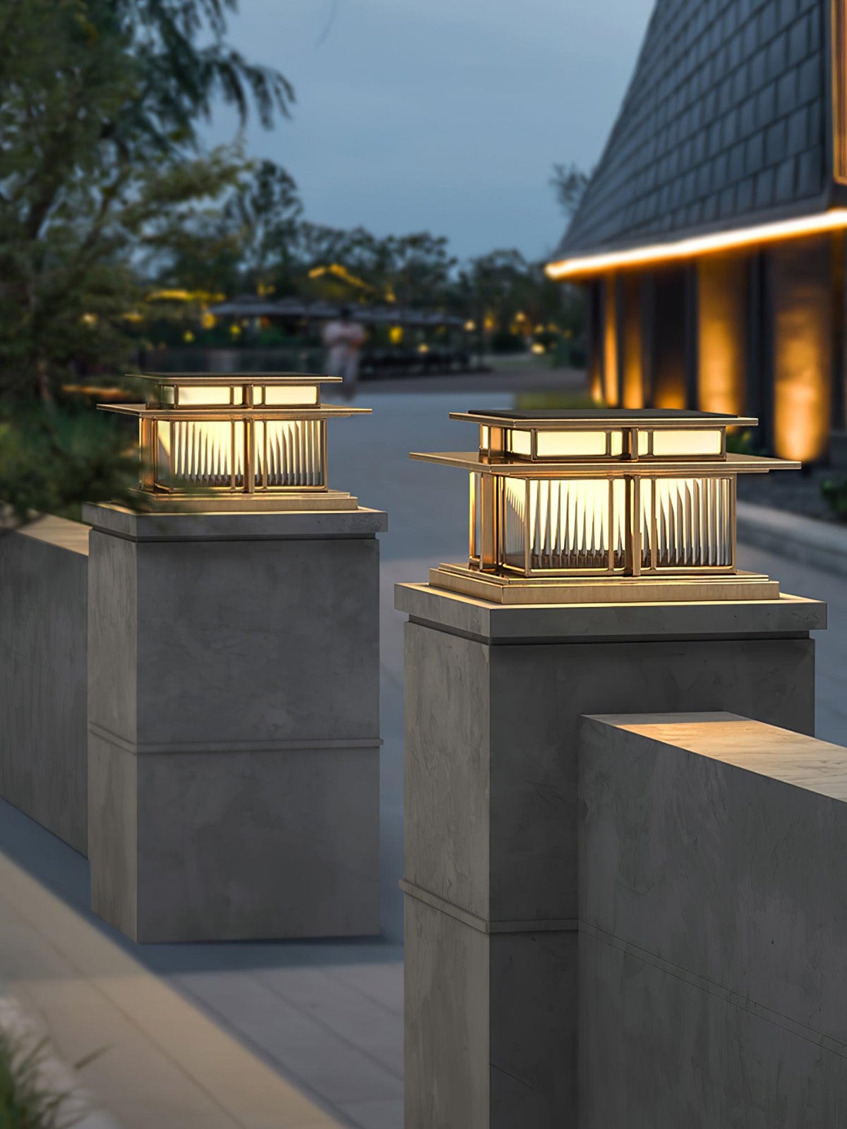 Two lit lanterns with a gold-toned frame stand atop stone posts at dusk, a different finish from this Clérachic pillar light's black frame.