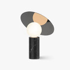 Clérachic tilting disc table lamp on a plain white background, its mirrored grey and gold panels angled above a black marble base.