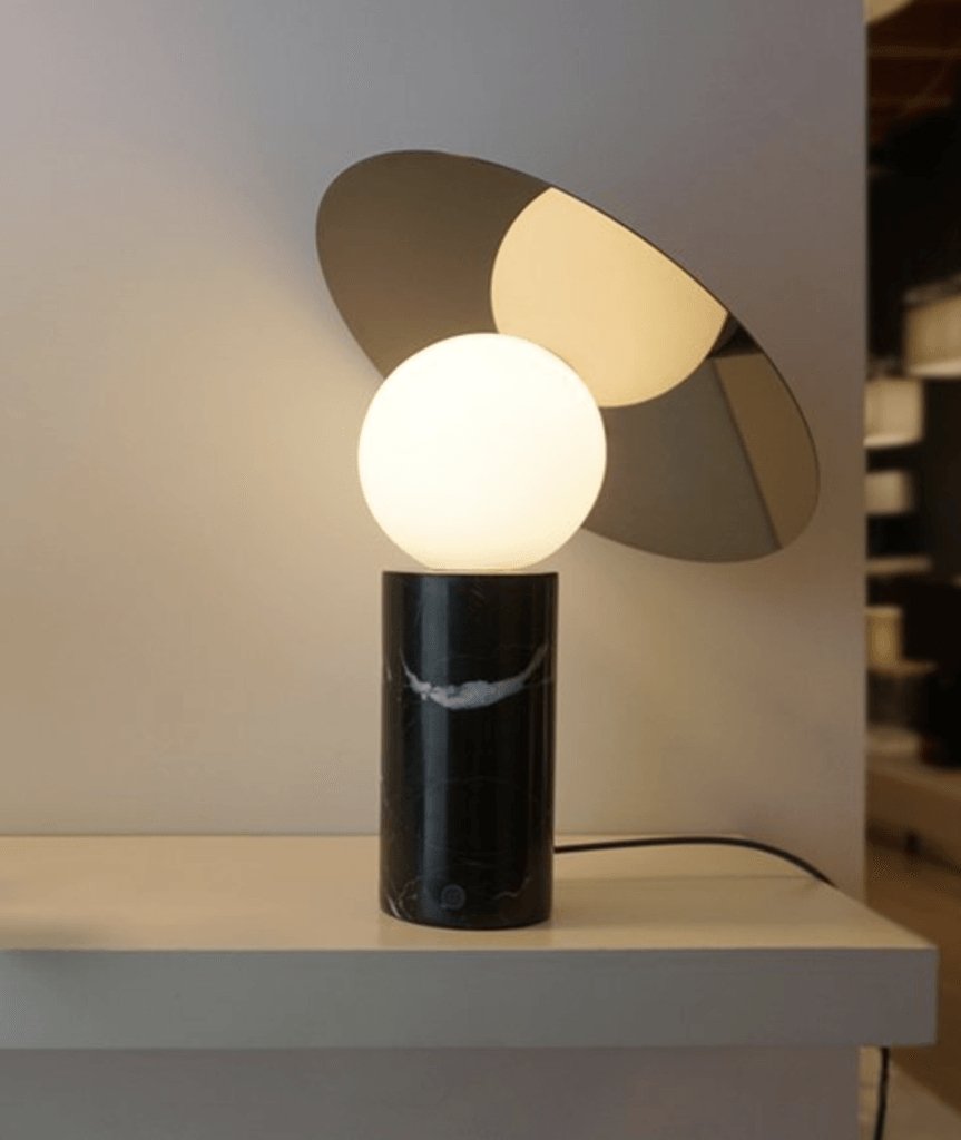 Against a grey wall with an abstract art print, the Clérachic tilting-disc lamp lights a showroom desk with no props beside it.