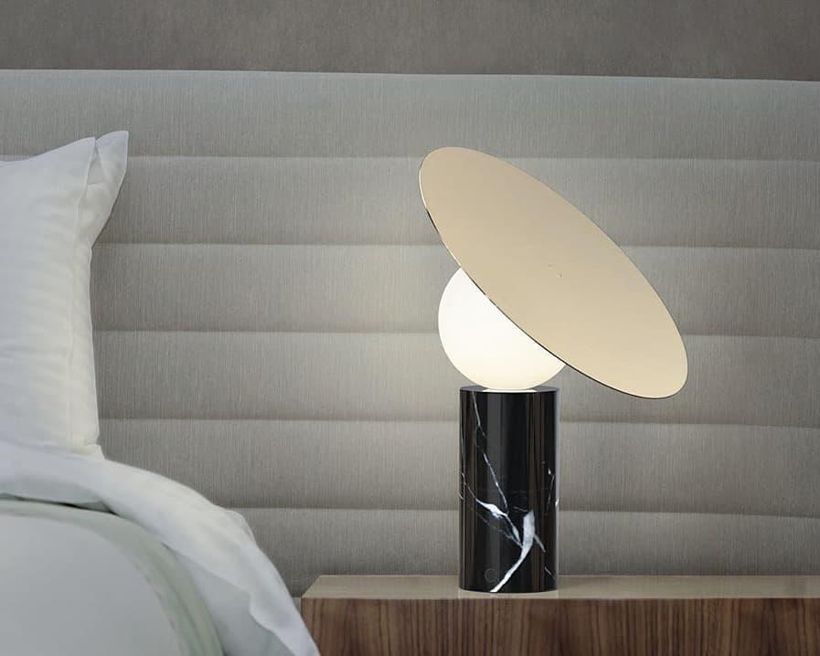 Below a pale grey channel-tufted headboard, the Clérachic disc lamp glows on a bedside table, its wood cabinet just in view.