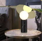 Clérachic lamp lit on a round grey marble-top side table near a sofa, a tropical leaf reaching into the edge of the frame.