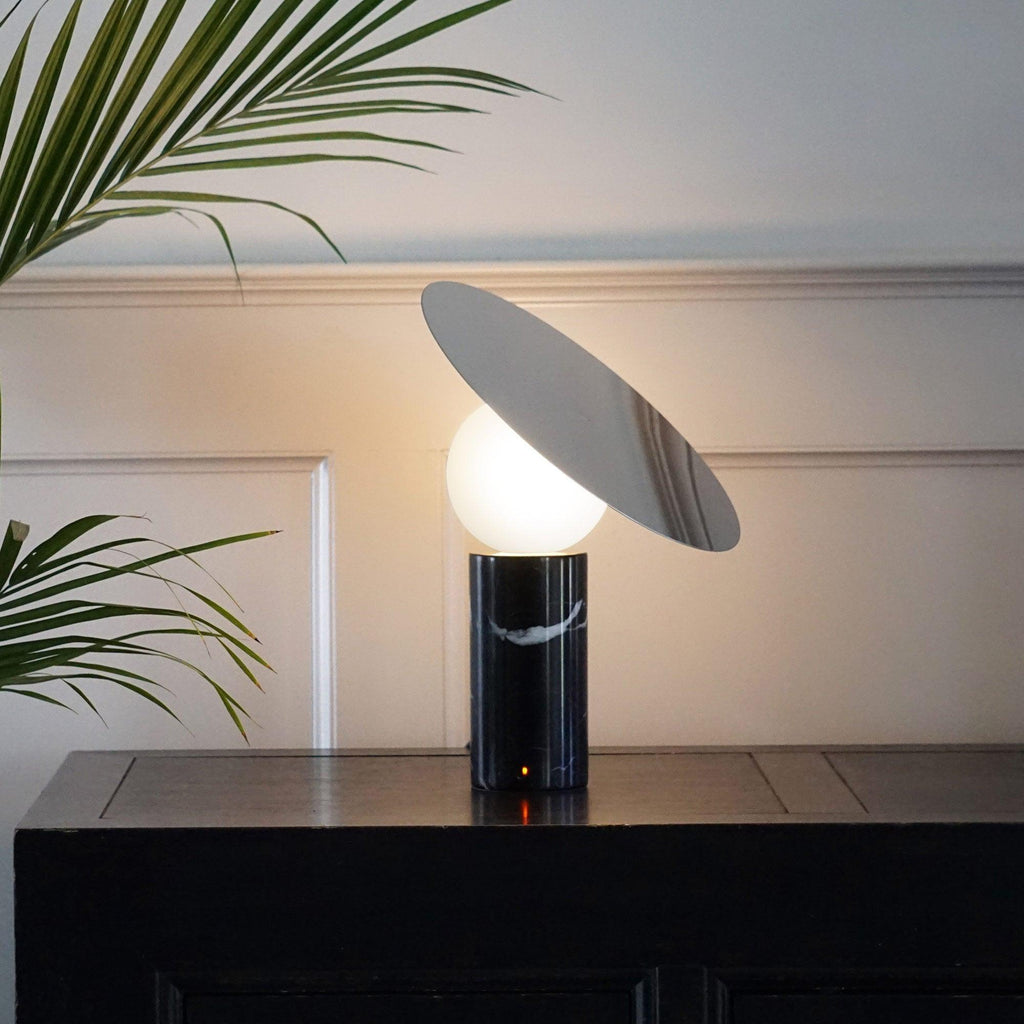 Clérachic lamp lit on a dark wood table against a panelled white wall, palm leaves reaching into frame beside the tilted disc.