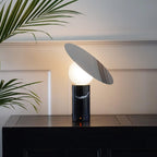 Clérachic lamp lit on a dark wood table against a panelled white wall, palm leaves reaching into frame beside the tilted disc.