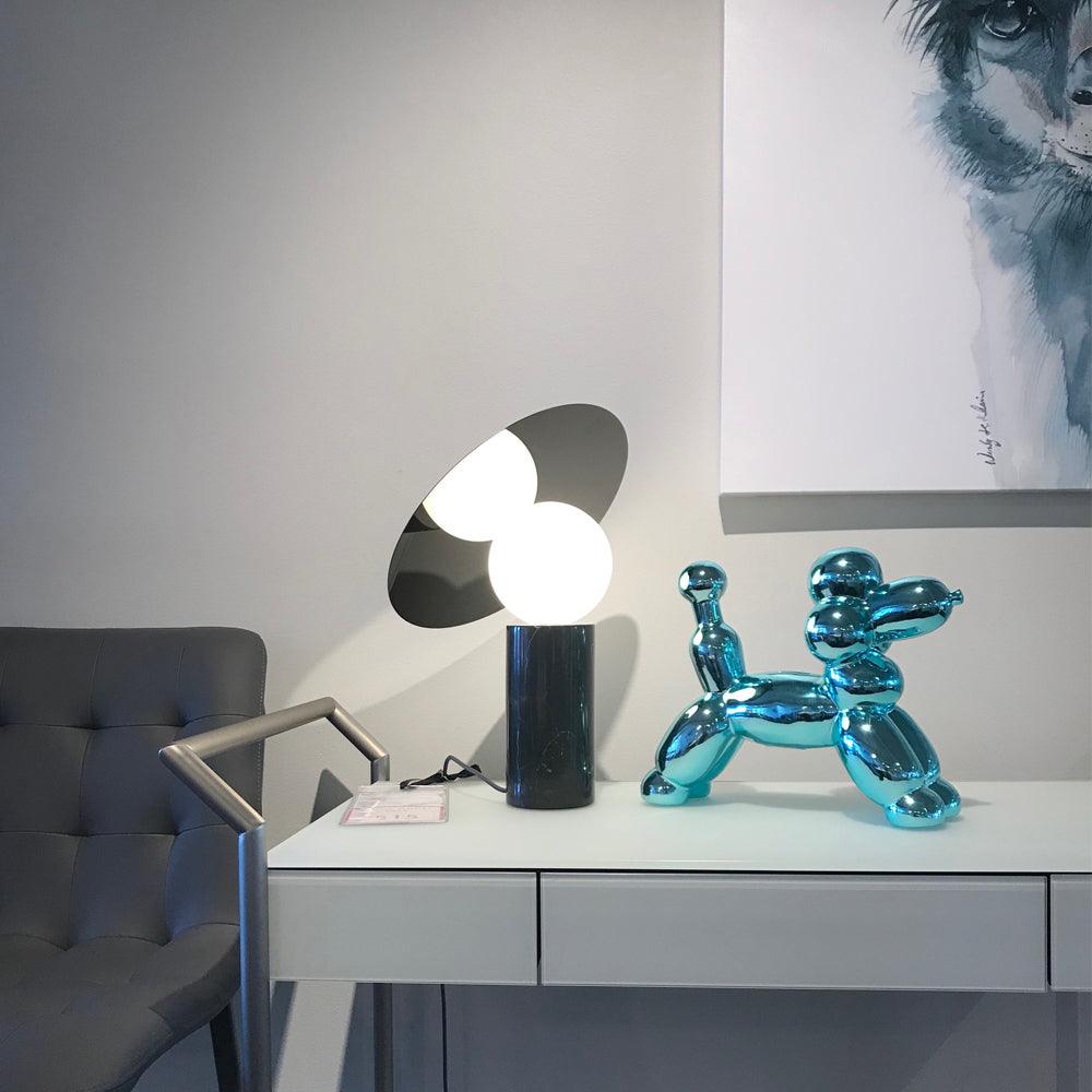 Clérachic tilting-disc lamp lit on a showroom desk with a price tag beside it, a chrome balloon-dog ornament and framed art in view.