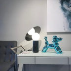 Clérachic tilting-disc lamp lit on a showroom desk with a price tag beside it, a chrome balloon-dog ornament and framed art in view.