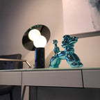 Beside a chrome balloon-dog ornament, the Clérachic disc lamp glows on a glass-topped showroom desk with grey upholstered chairs nearby.