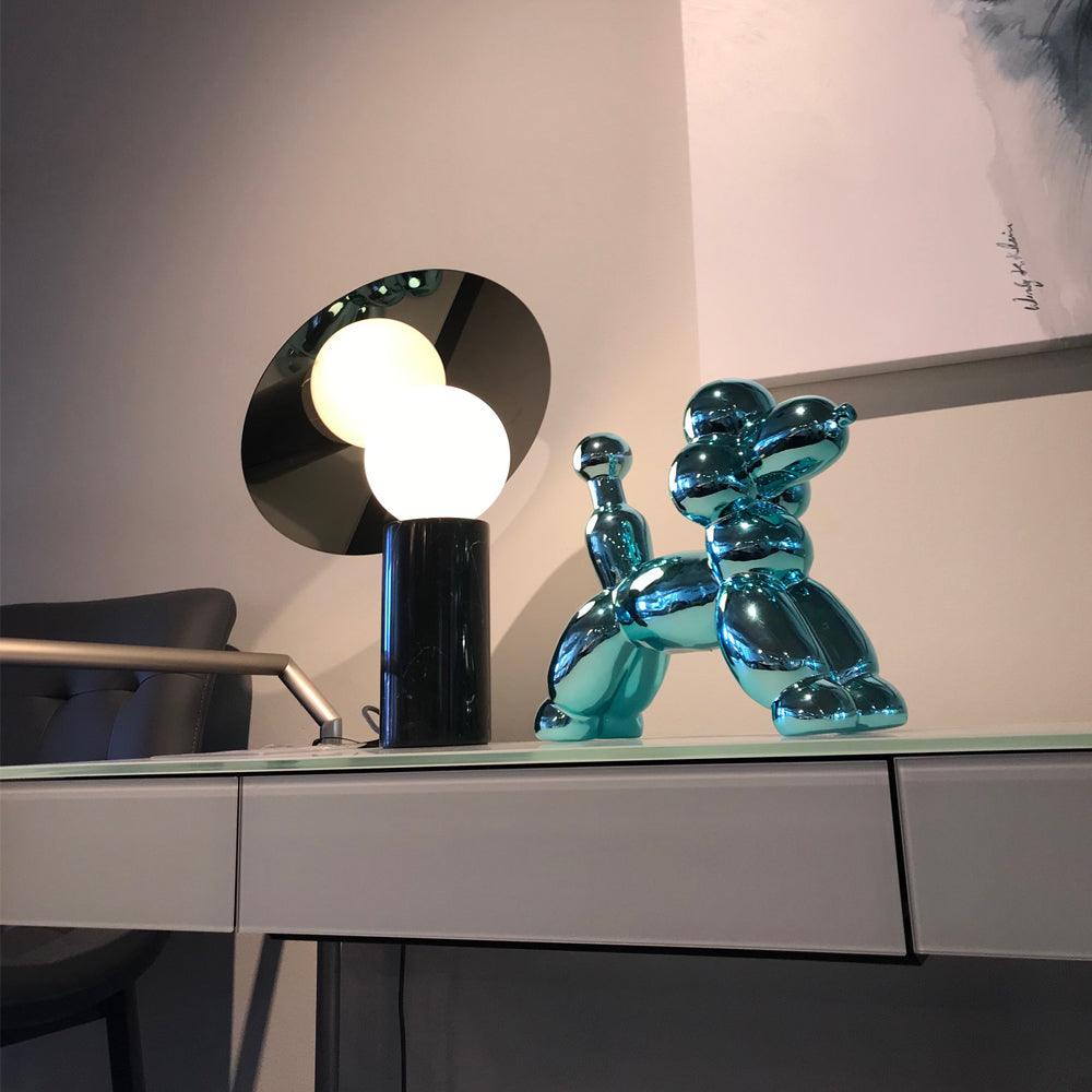 Beside a chrome balloon-dog ornament, the Clérachic disc lamp glows on a glass-topped showroom desk with grey upholstered chairs nearby.