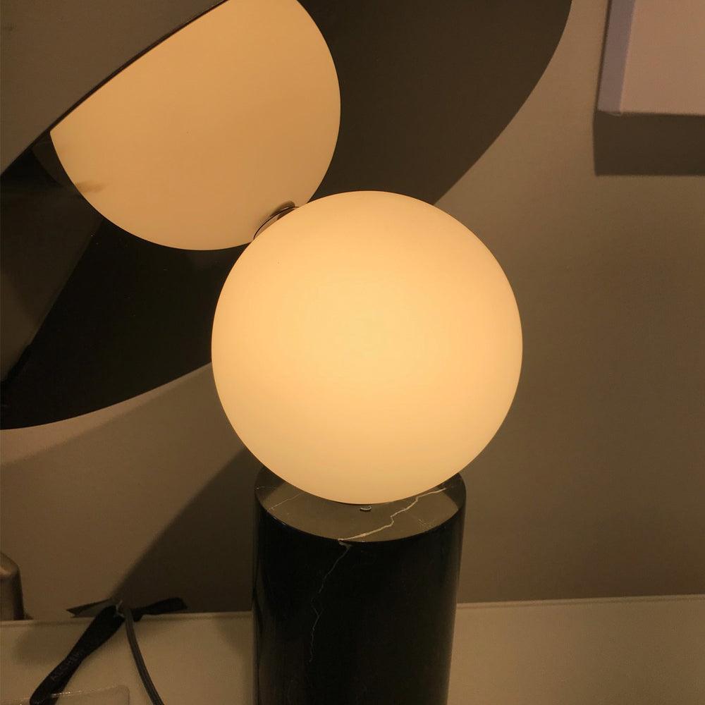 Extreme close-up of the Clérachic lamp's gold-toned panel and lit frosted glass globe, a dim interior blurred behind it.