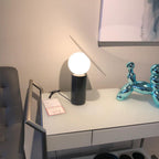Clérachic tilting-disc lamp lit on a showroom desk with a price tag visible, a grey upholstered chair beside the table.