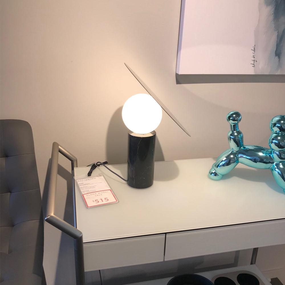 Clérachic tilting-disc lamp lit on a showroom desk with a price tag visible, a grey upholstered chair beside the table.