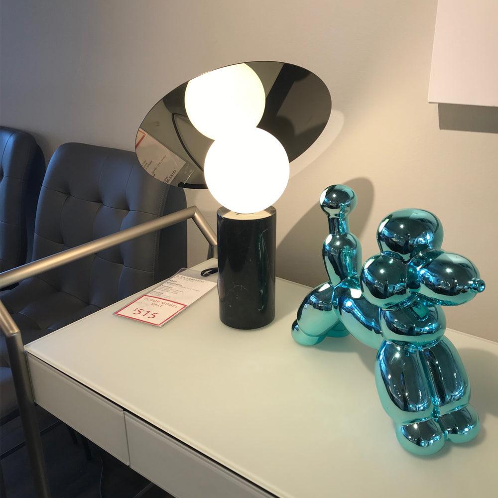 Between tufted grey chairs, the Clérachic disc lamp glows on a showroom desk beside a price tag and chrome balloon-dog ornament.
