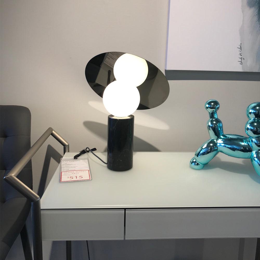 Tufted grey chairs frame the scene as the Clérachic disc lamp glows on a showroom desk beside a chrome balloon-dog ornament.