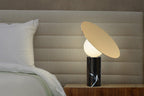 Against a pale grey headboard, the Clérachic disc lamp glows on a bedside table, its black marble base set on a dark wood cabinet.