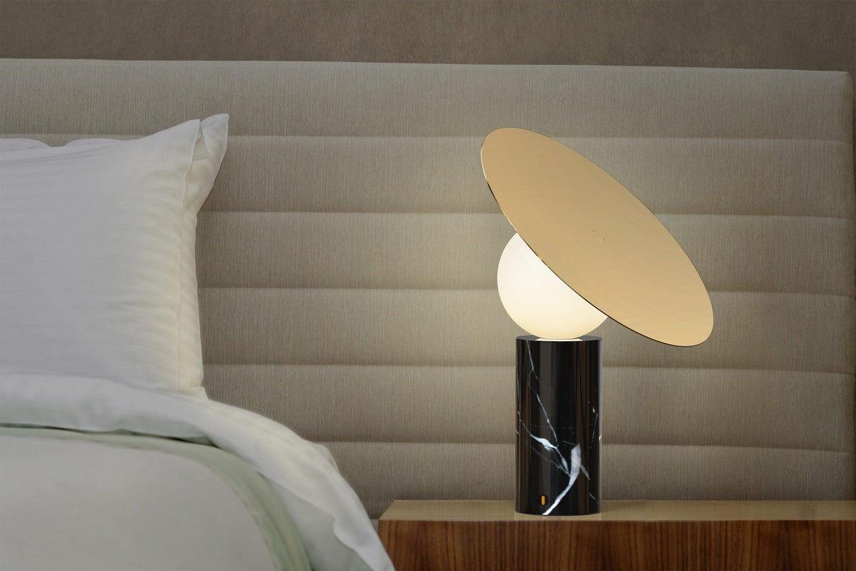 Against a pale grey headboard, the Clérachic disc lamp glows on a bedside table, its black marble base set on a dark wood cabinet.