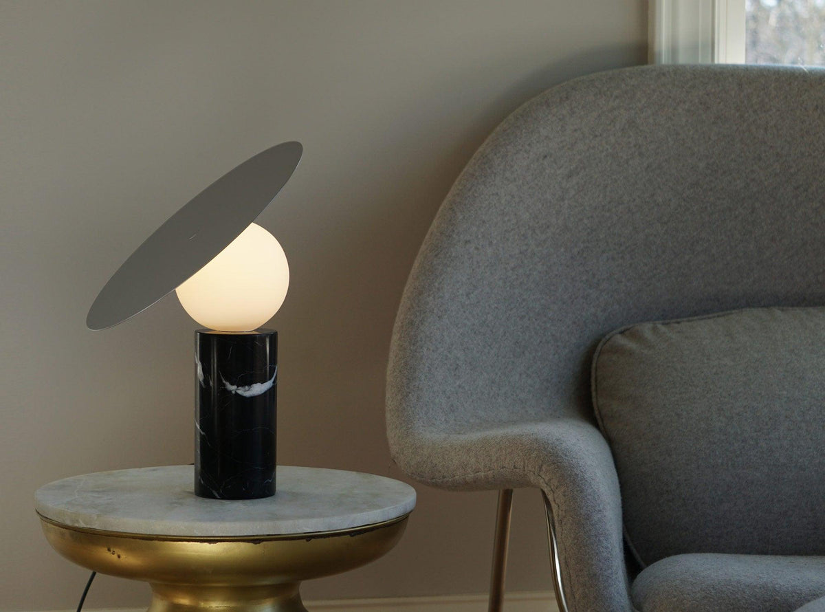 A grey bouclé armchair fills the background as the Clérachic tilting-disc lamp lights a small brass-legged marble side table.