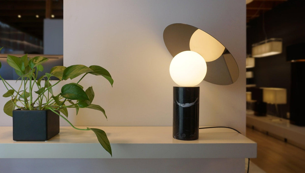 The Clérachic lamp glows on a console shelf beside a potted plant, a blurred store interior with pendant lights behind it.