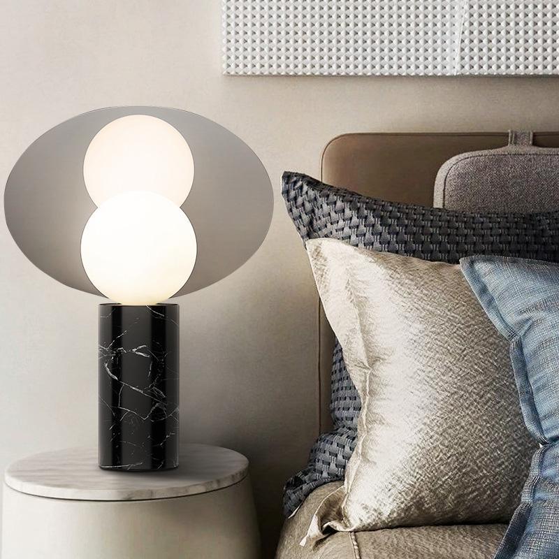 Clérachic disc lamp lit on a round white marble-top side table beside a bed with a grey woven headboard and pillows.