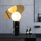 Clérachic tilting-disc lamp lit in a bathroom against a textured concrete wall, argan oil bottles standing near its marble base.