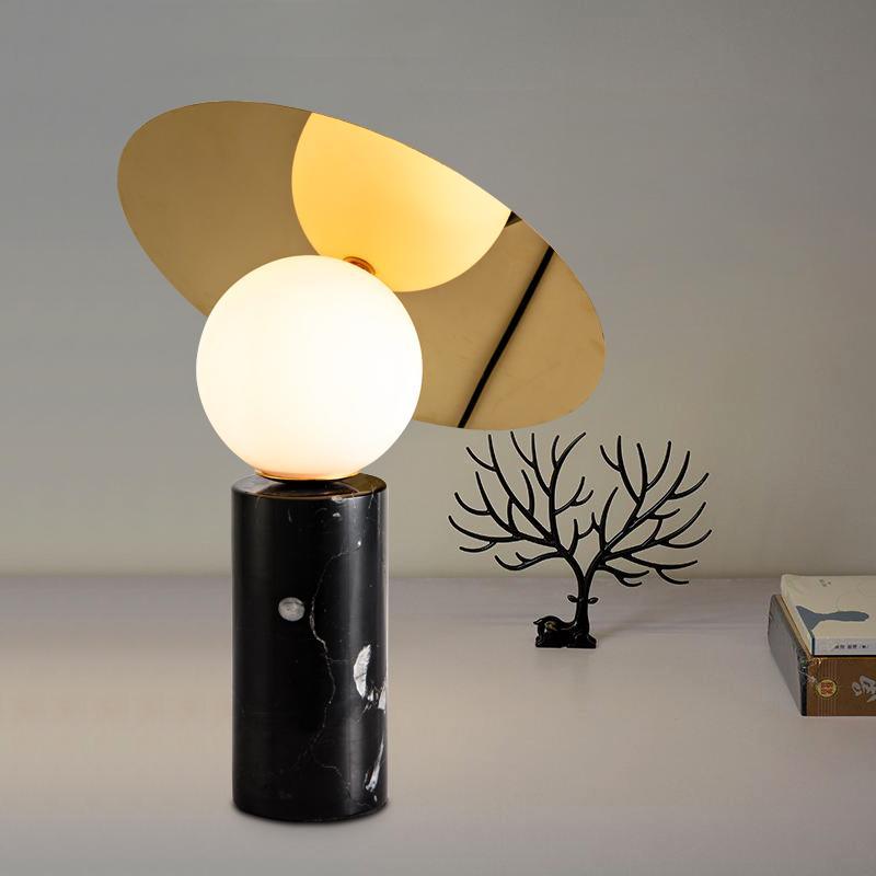 Clérachic table lamp lit on a wood-topped table against a beige wall, a small black coral-branch ornament standing beside its marble base.