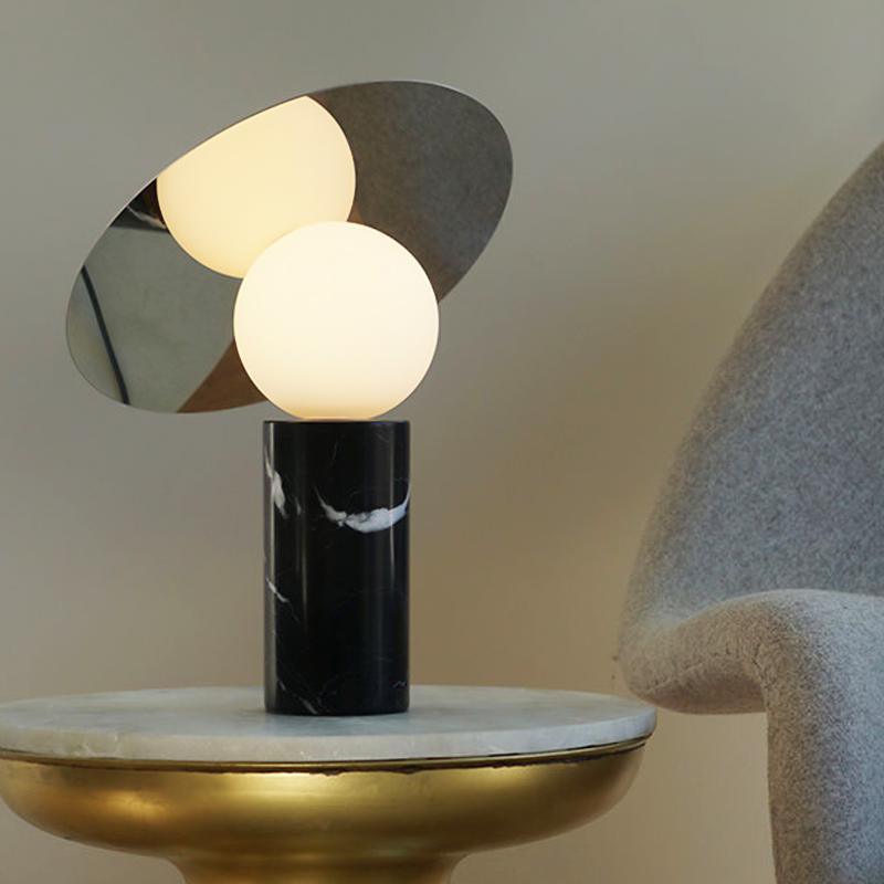 Clérachic tilting-disc lamp lit on a round marble-top side table beside a grey bouclé armchair, its black marble base in the foreground.