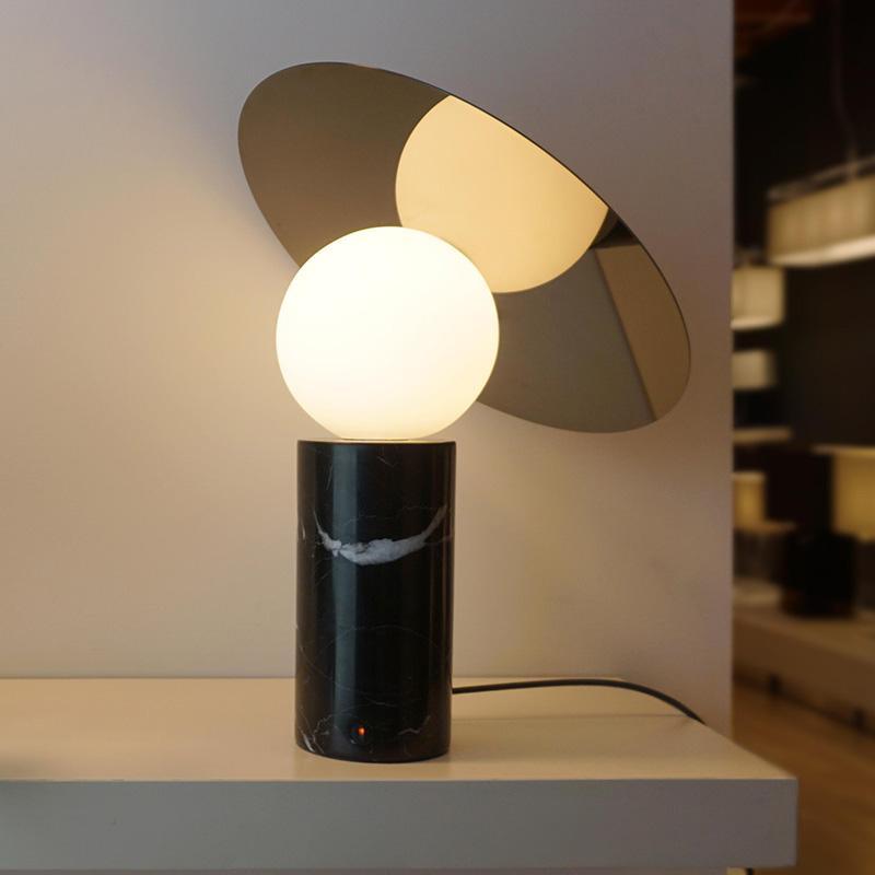 The Clérachic disc lamp glows warm on a white shelf edge, its cylindrical black marble base set against a blurred room with pendant lights.