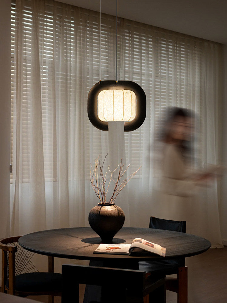 Before sheer curtains, this black, closed-loop Clérachic pendant glows above a round table set with a vase and an open book.