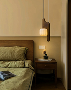 In a bedroom with olive bedding, this dark wood, bent-style Clérachic pendant glows beside a wood nightstand.